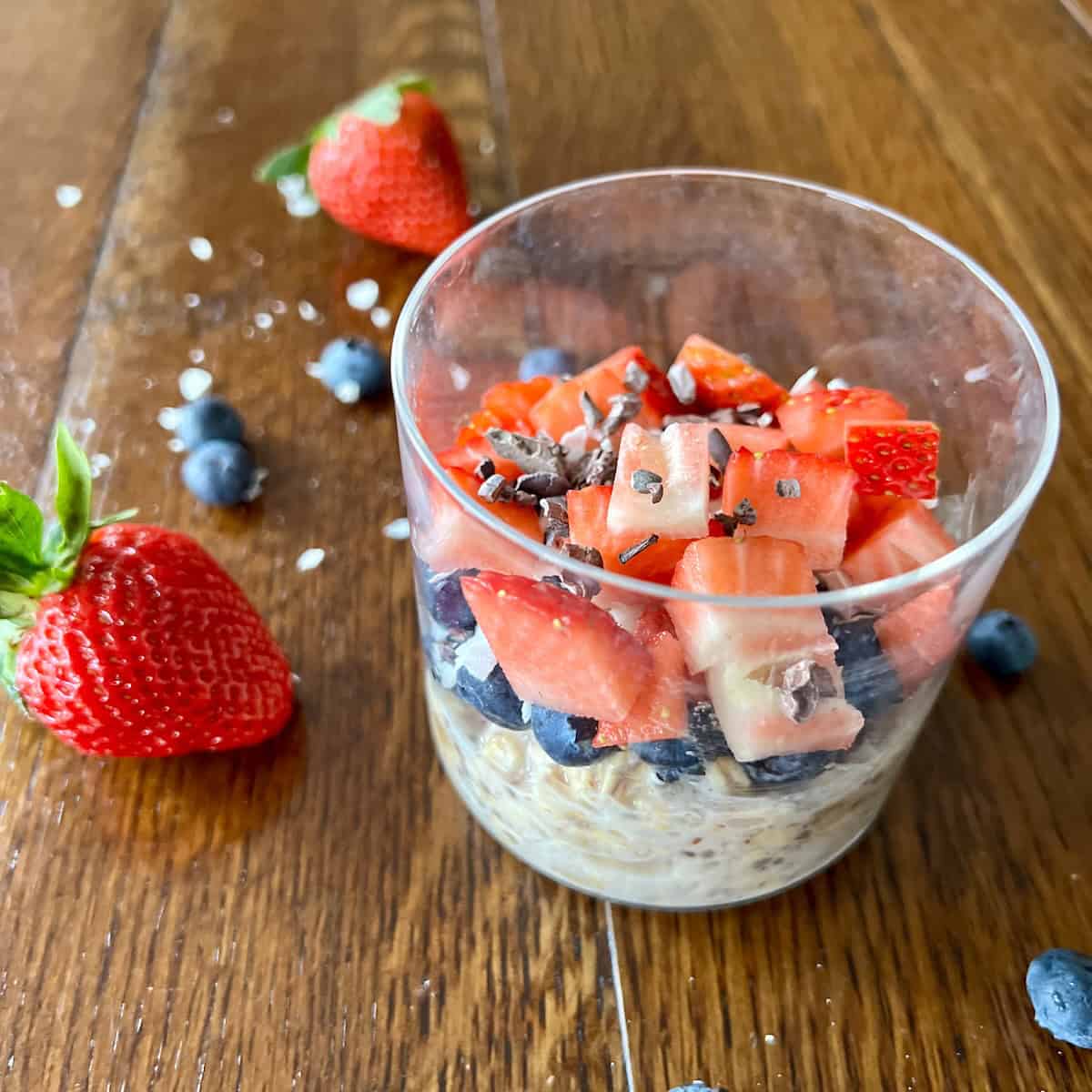 a round glass containing classic overnight oats topped with blueberries, strawberries, shredded coconut, and cacao nibs