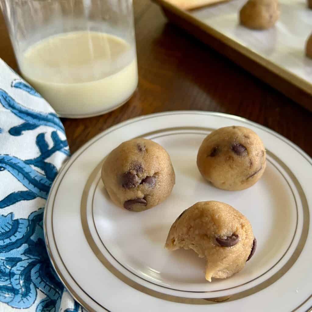 small plate of three banana bread cookie dough freezer bites with a bite taken out of one of them and a glass of milk and baking sheet in the background