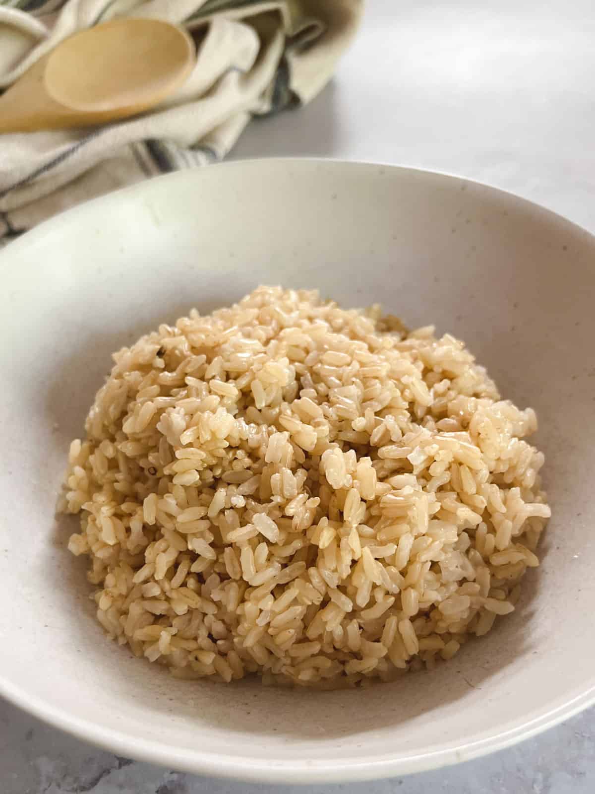 close up of bowl of cooked brown rice with towel and wooden spoon in the background