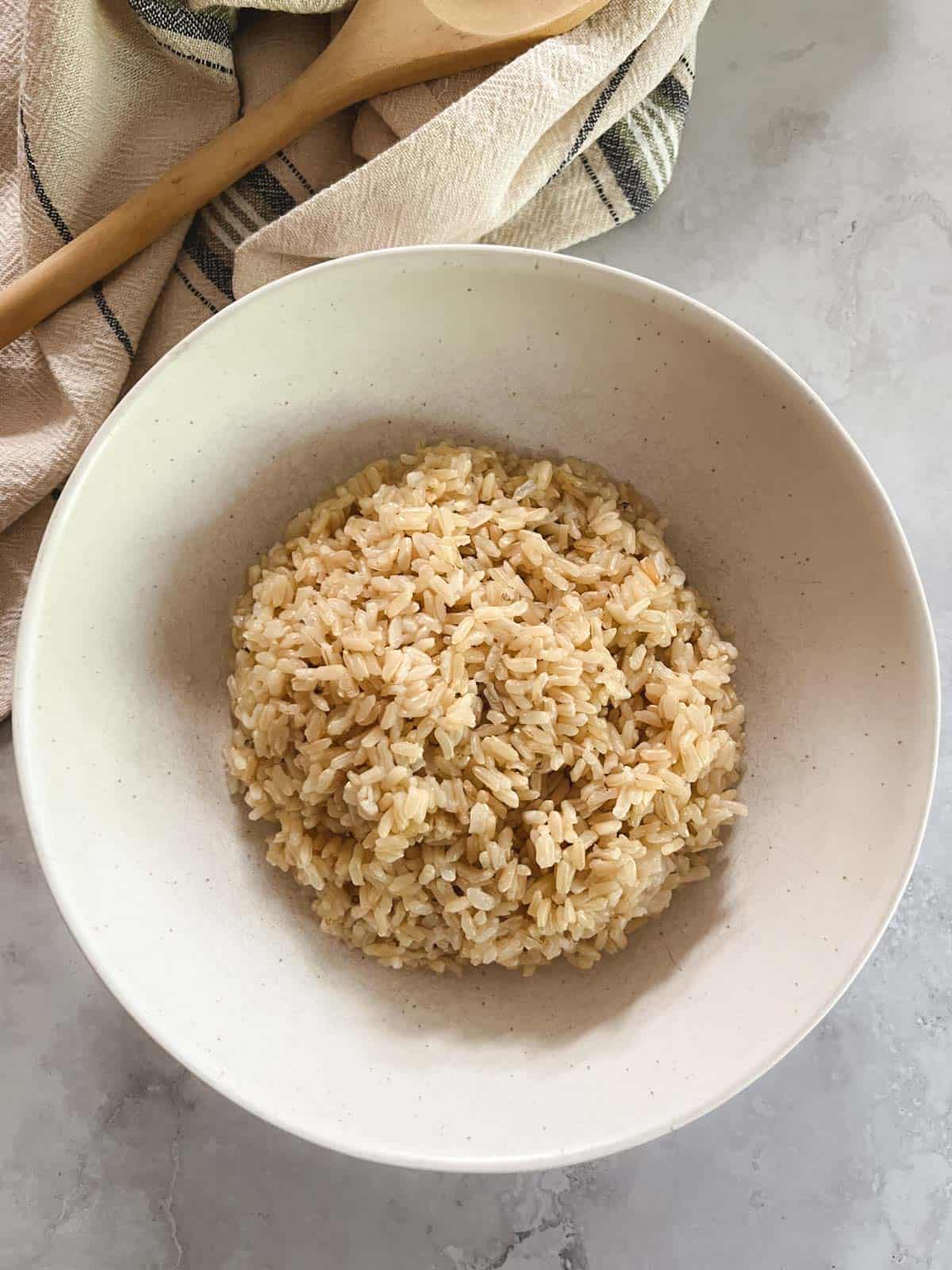 overhead view of bowl of cooked brown rice with a towel and wooden spoon