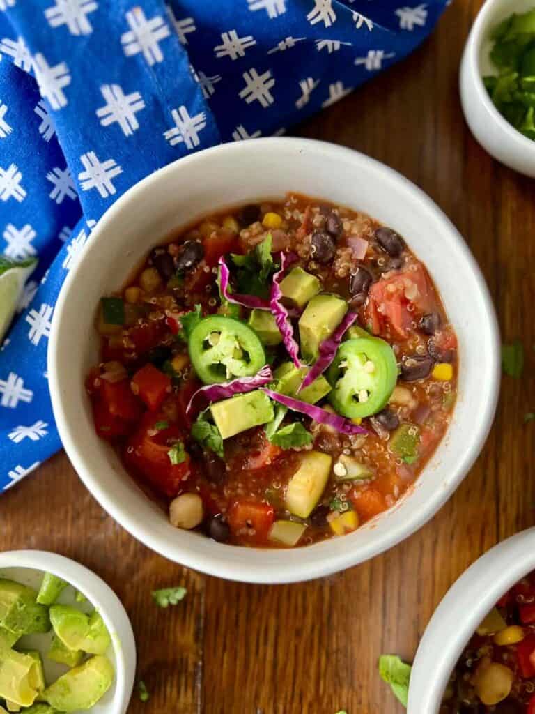 bowl of zesty quinoa chili on a wood table surrounded by blue napkin and small bowls of diced avocado and cilantro