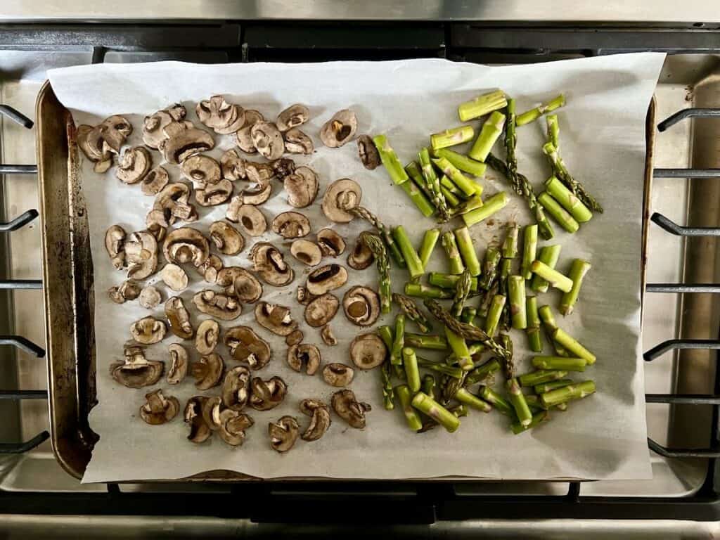 baking sheet lined with parchment paper with cooked mushrooms on left side and asparagus chunks on the right side 