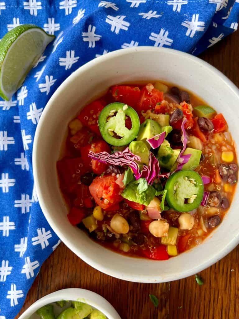 overhead close up of a round bowl of zesty quinoa chili topped with diced avocado, shredded purple cabbage, and sliced jalapeños