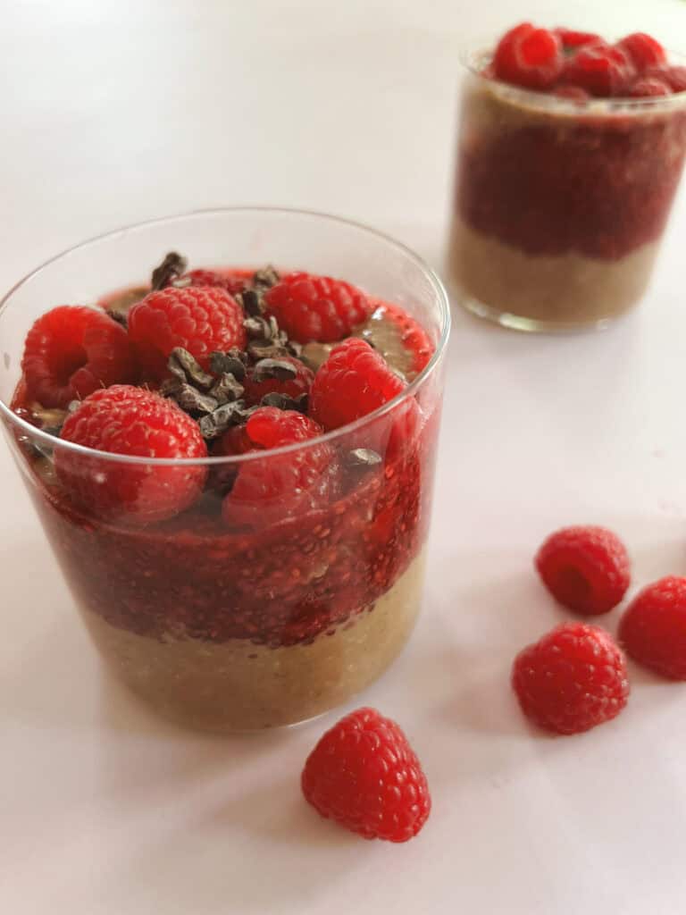 two glasses of creamy layered cocoa raspberry overnight oats with scattered raspberries on the side