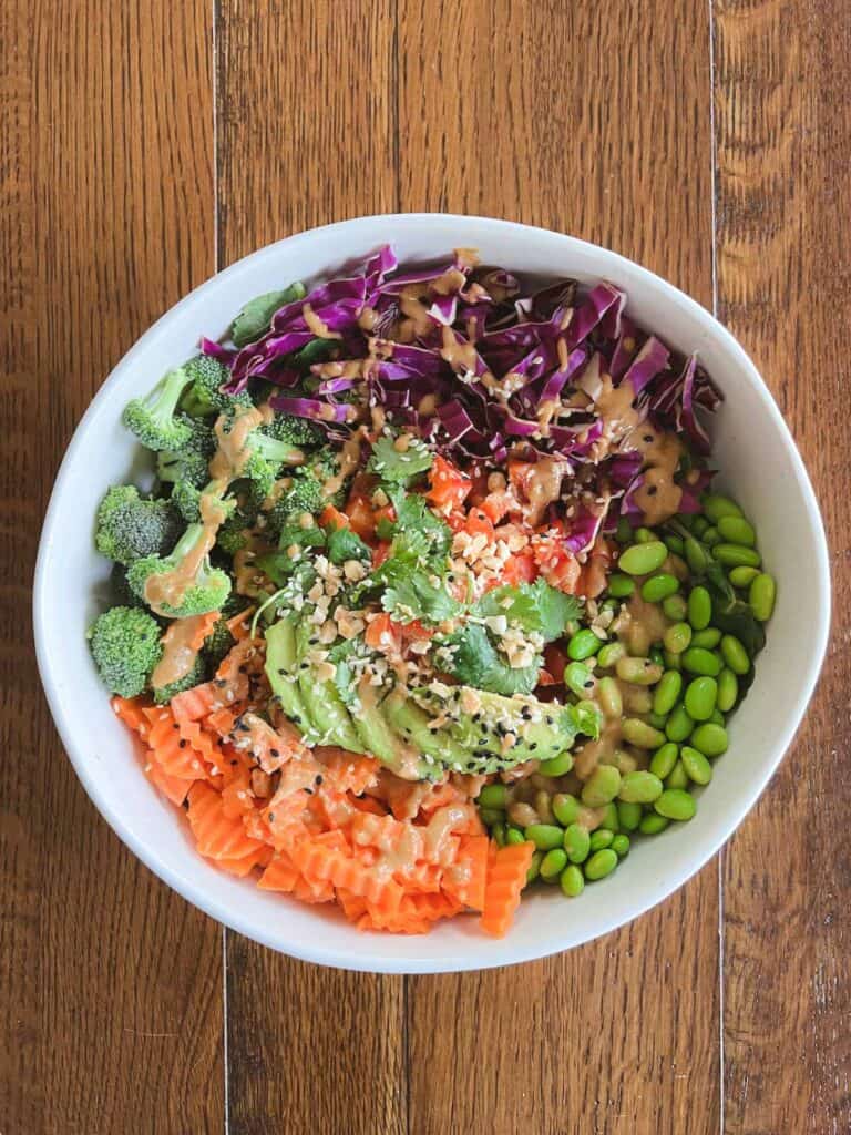 overhead of rainbow crunch edamame salad in a bowl topped with dressing, sliced avocado, cilantro, crushed peanuts, and sesame seeds