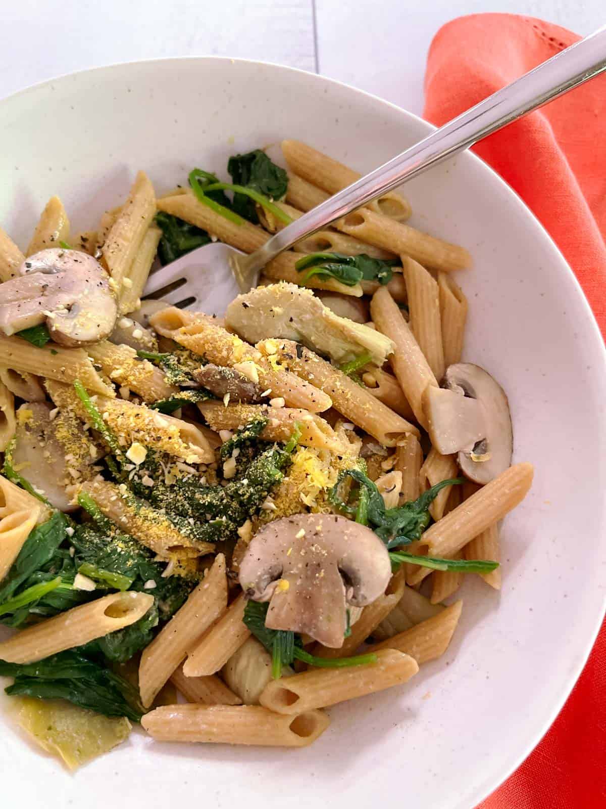 close up of a bowl of spinach, mushroom, and artichoke pasta with a fork