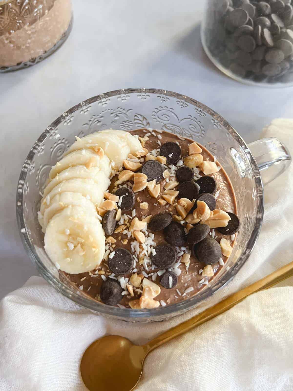 glass mug containing creamy blended chocolate peanut butter overnight oats with toppings, with jar of chocolate chips and additional overnight oats serving in the background