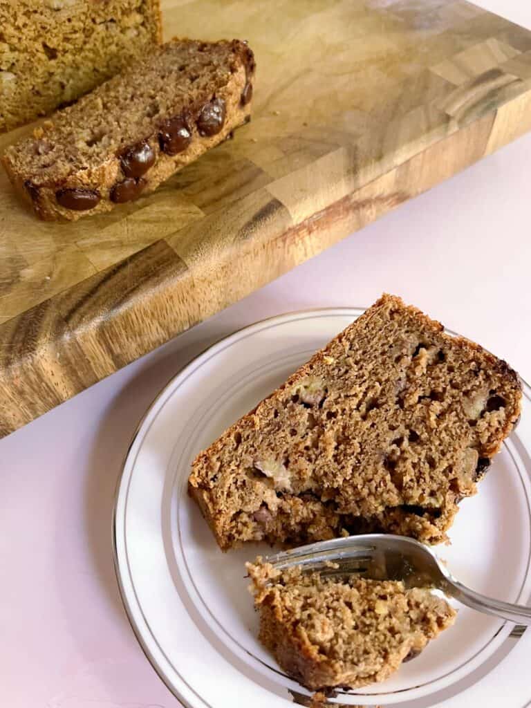 slice of banana bread on a plate with a fork with a cutting board with sliced banana bread in the background