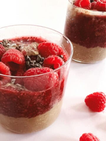 two glasses of creamy layered cocoa raspberry overnight oats with scattered raspberries