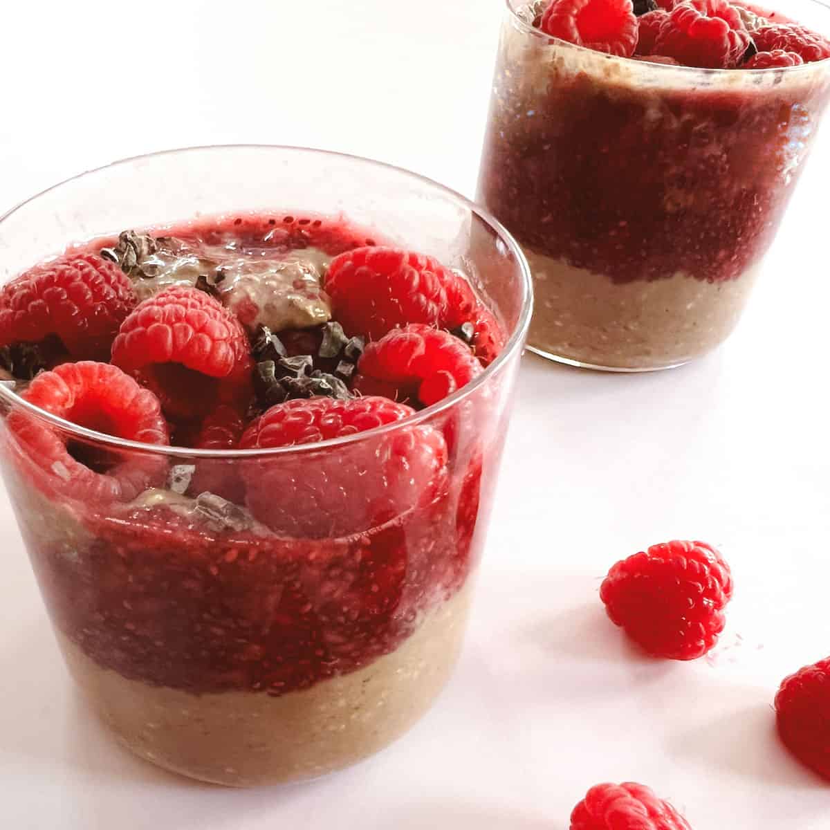 two glasses of creamy layered cocoa raspberry overnight oats with scattered raspberries