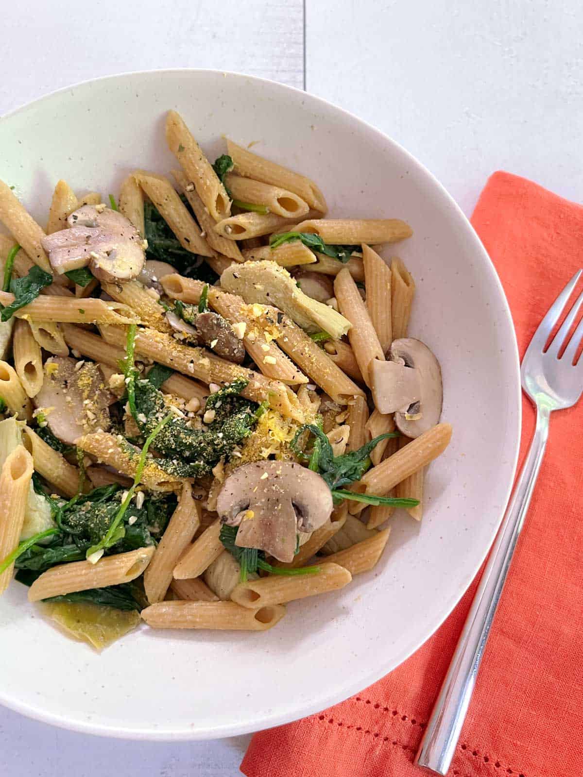 bowl of spinach, mushroom, and artichoke pasta with a fork and napkin on the right side