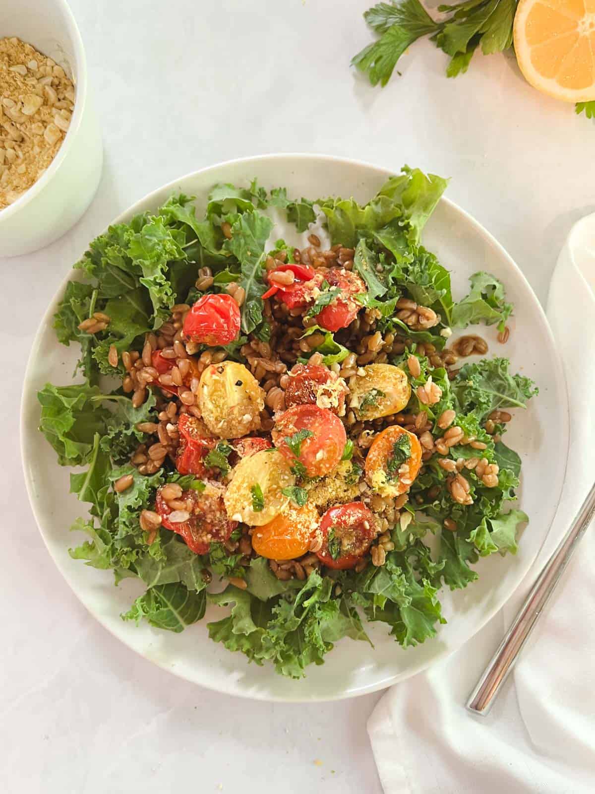 plate of farro and kale salad with blistered tomatoes with a bowl of vegan Parmesan and a lemon in the background