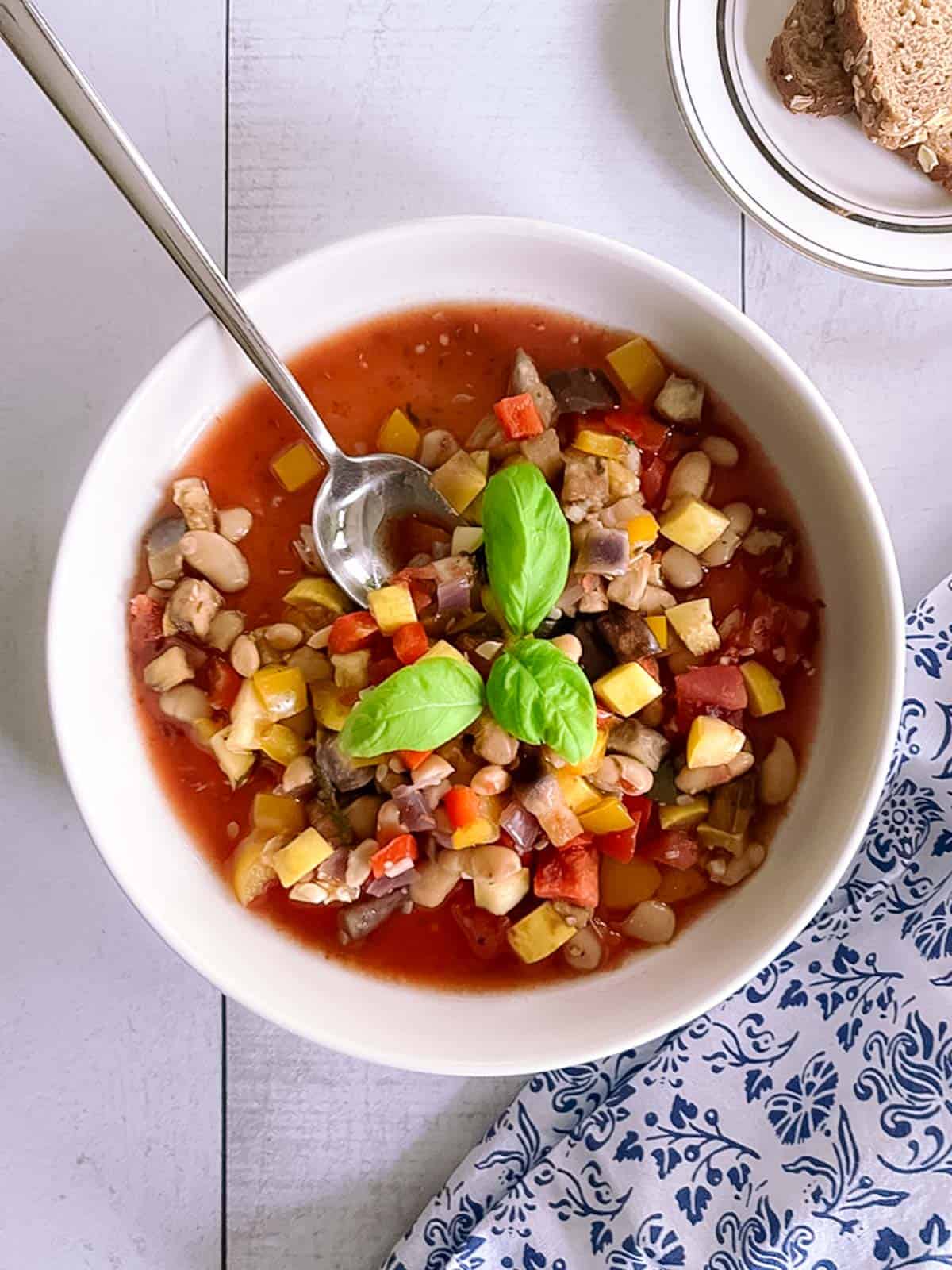 overhead view of a bowl of slow cooker ratatouille with white beans with a spoon in it; decorative napkin and plate of sliced bread to the side