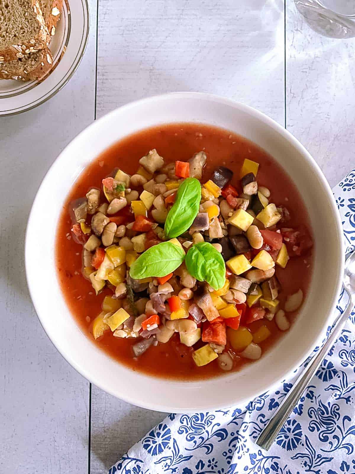 bowl of slow cooker ratatouille with white beans on a white wood table with a napkin and a plate of sliced bread