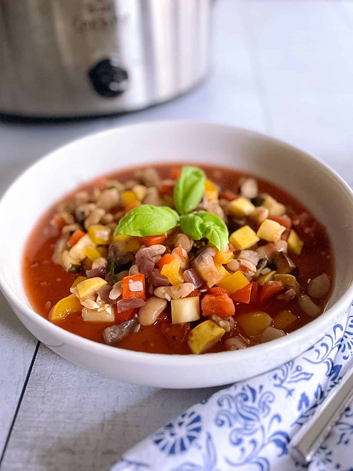 closeup of a bowl of slow cooker ratatouille with white beans and a napkin with a blurred crock pot in the background