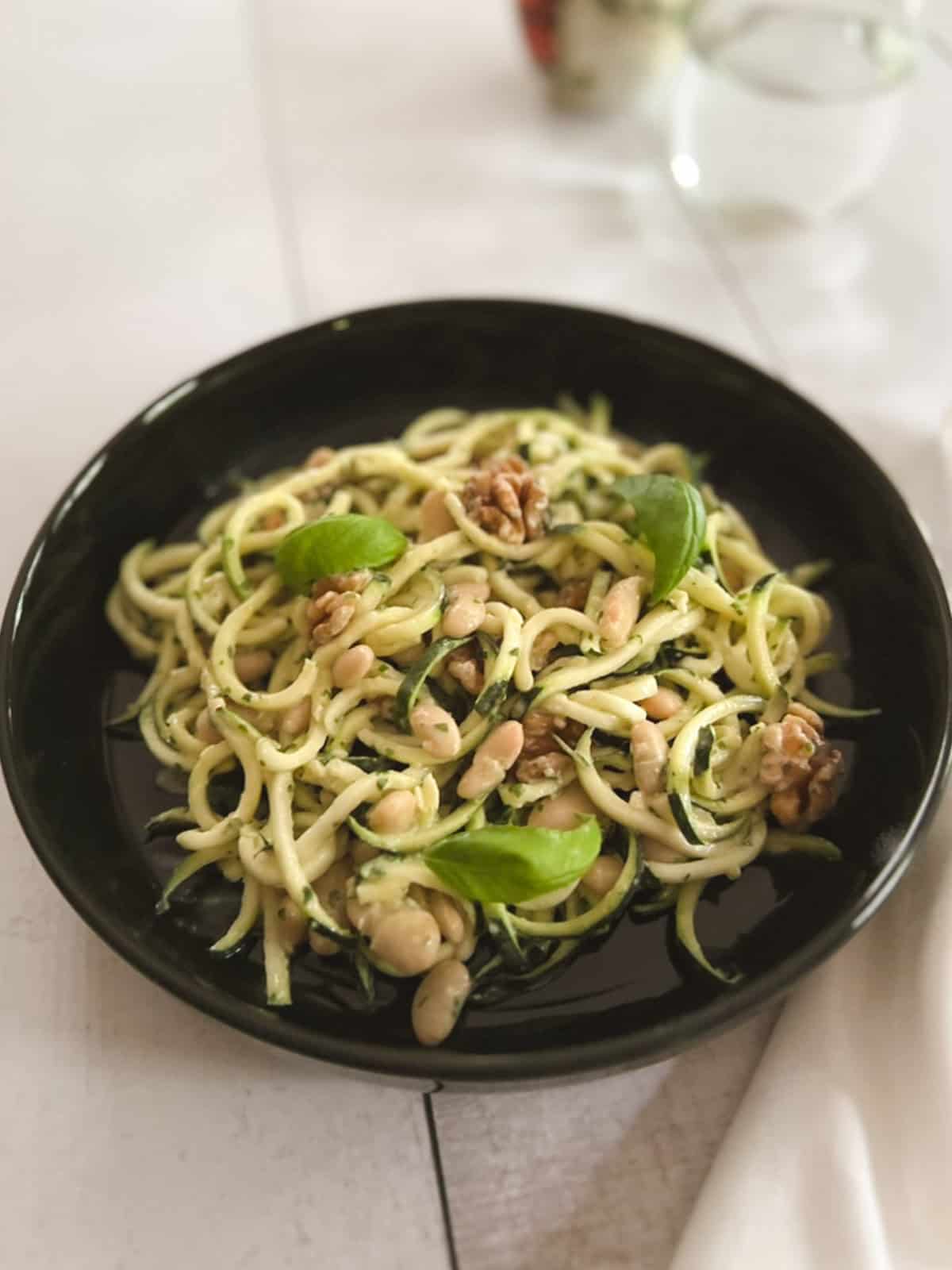 closeup of a bowl of zucchini noodles with tahini basil sauce with drinking glass and a vase in the background