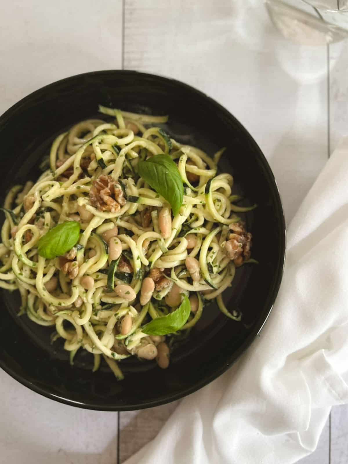 overhead of a bowl of zucchini noodles with tahini basil sauce with a glass in the background and a napkin to the side