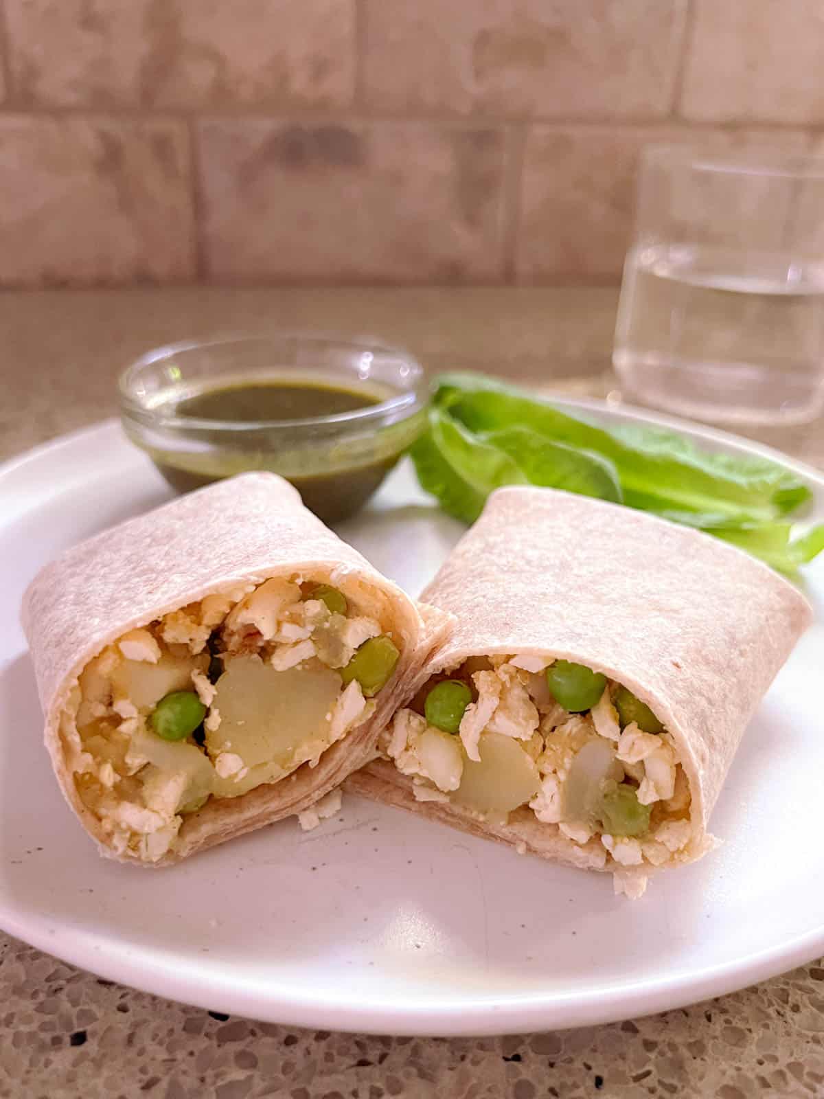 closeup of plate holding sliced potato and pea samosa wrap, bowl of mint chutney, and lettuce garnish