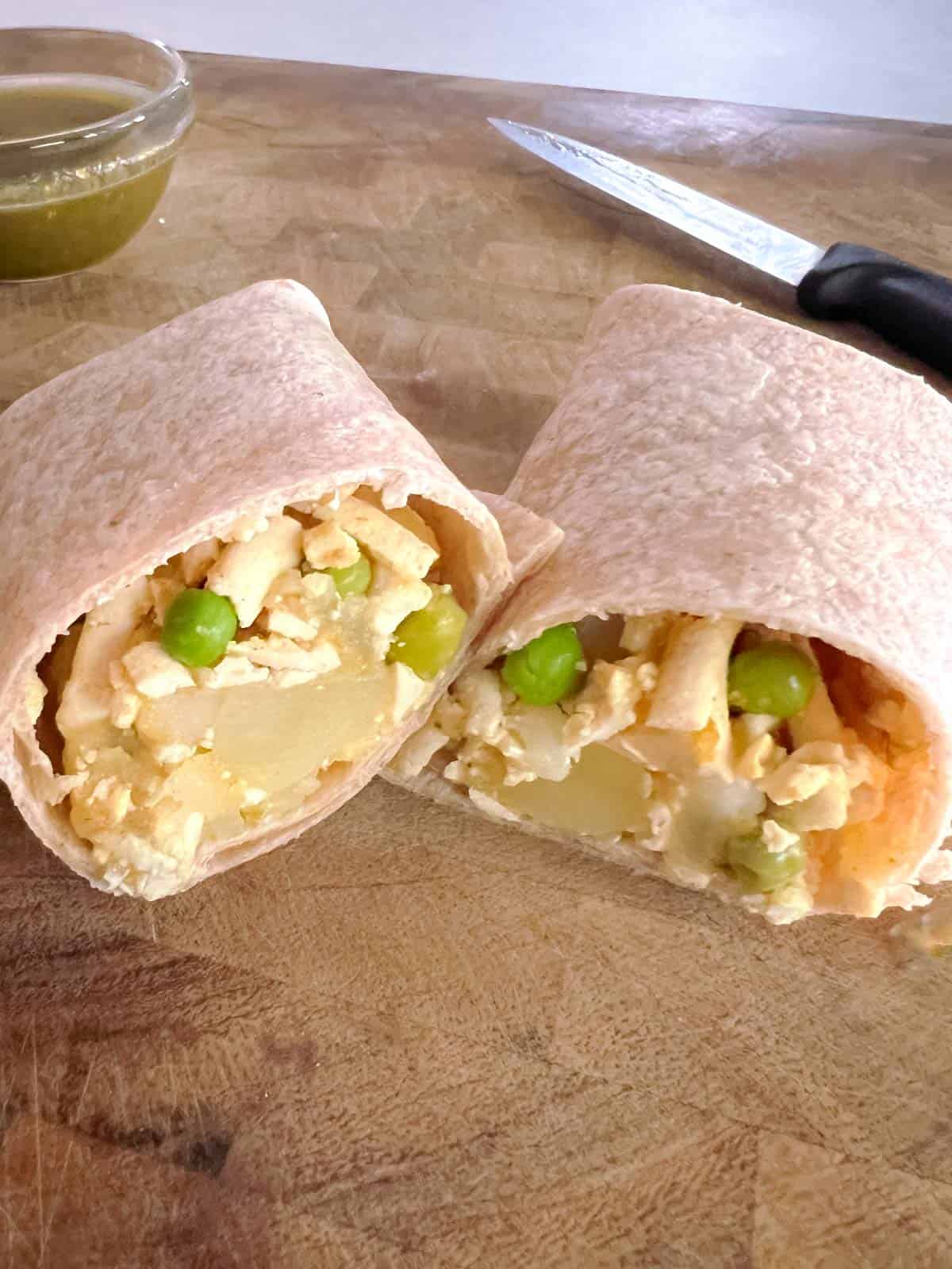 closeup of sliced potato and pea samosa wrap on a cutting board with knife and bowl of mint chutney in background