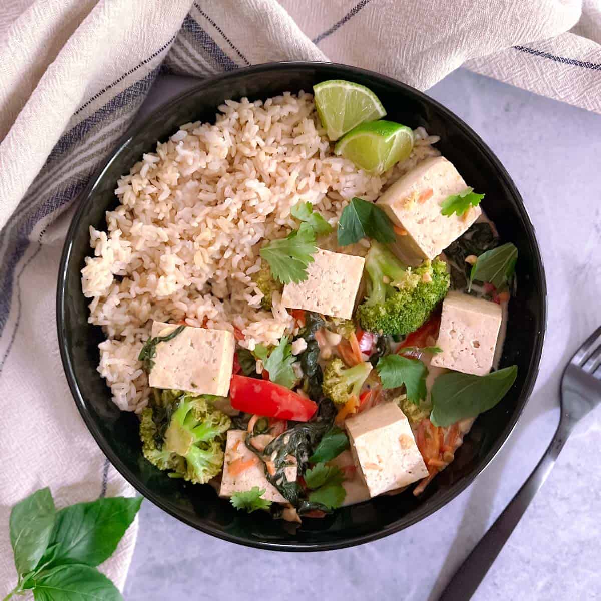 plate of easy vegan green curry with tofu and vegetables with a striped towel and basil leaves