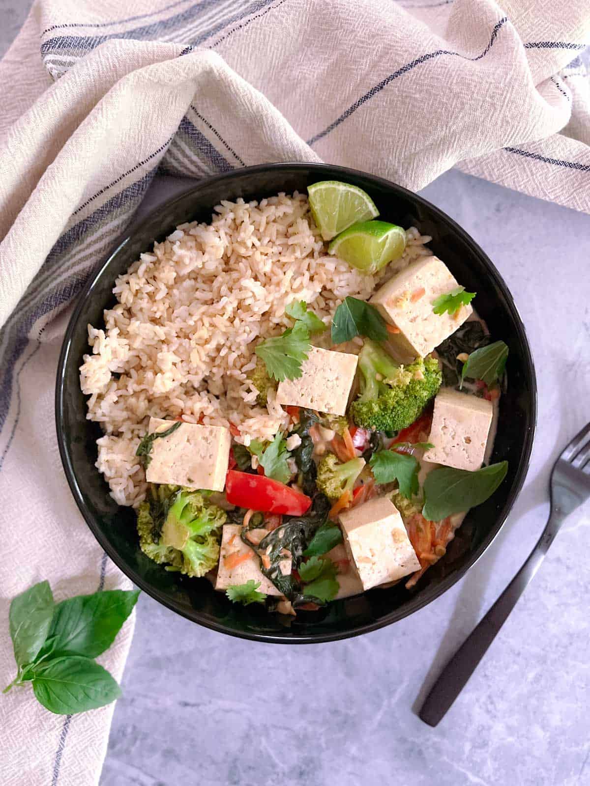 bowl of easy vegan green curry with tofu and vegetables with a fork and Thai basil garnish