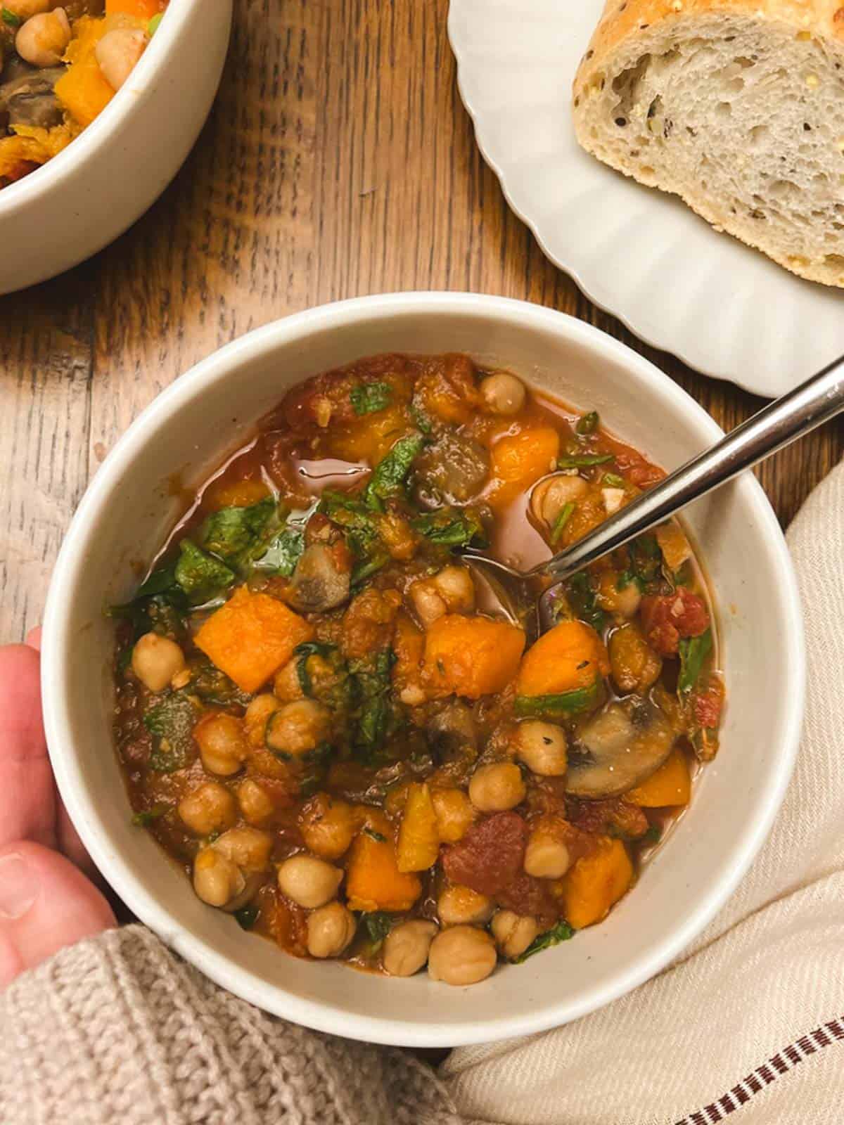 bowl of slow cooker fall harvest ratatouille held in a hand, with a plate of bread in the background 