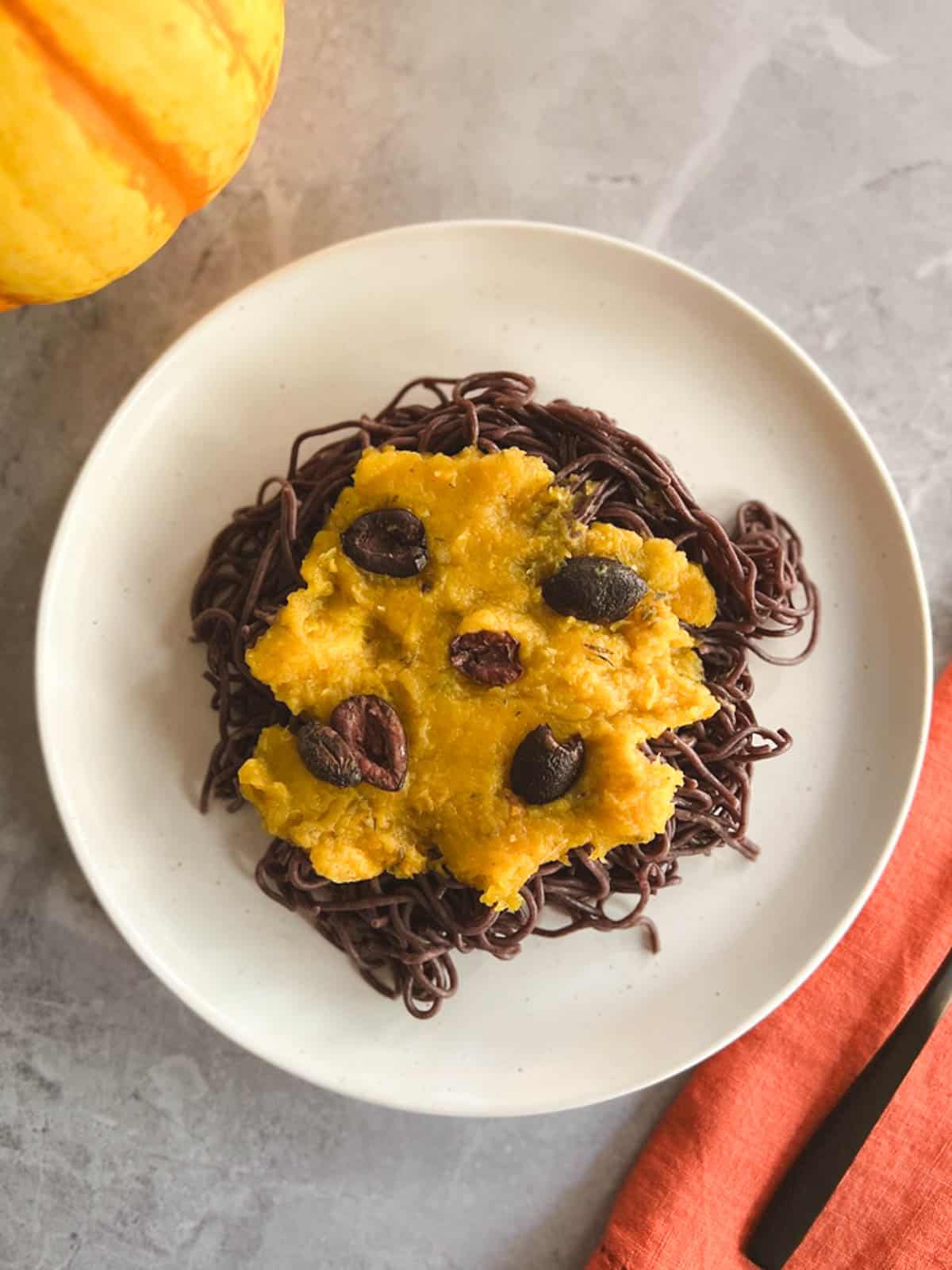 a serving of vegan halloween pasta garnished with roasted olives on a plate with a pumpkin in the background