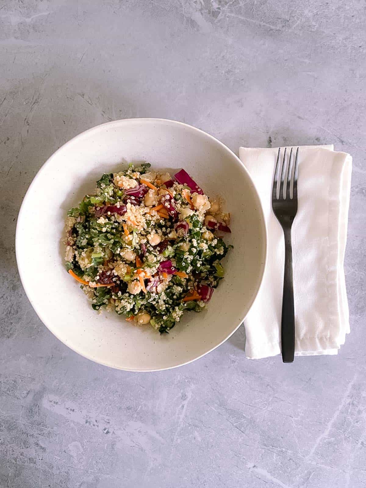 overhead view of a white bowl containing a serving of autumn quinoa salad with a black fork on a white napkin to the right