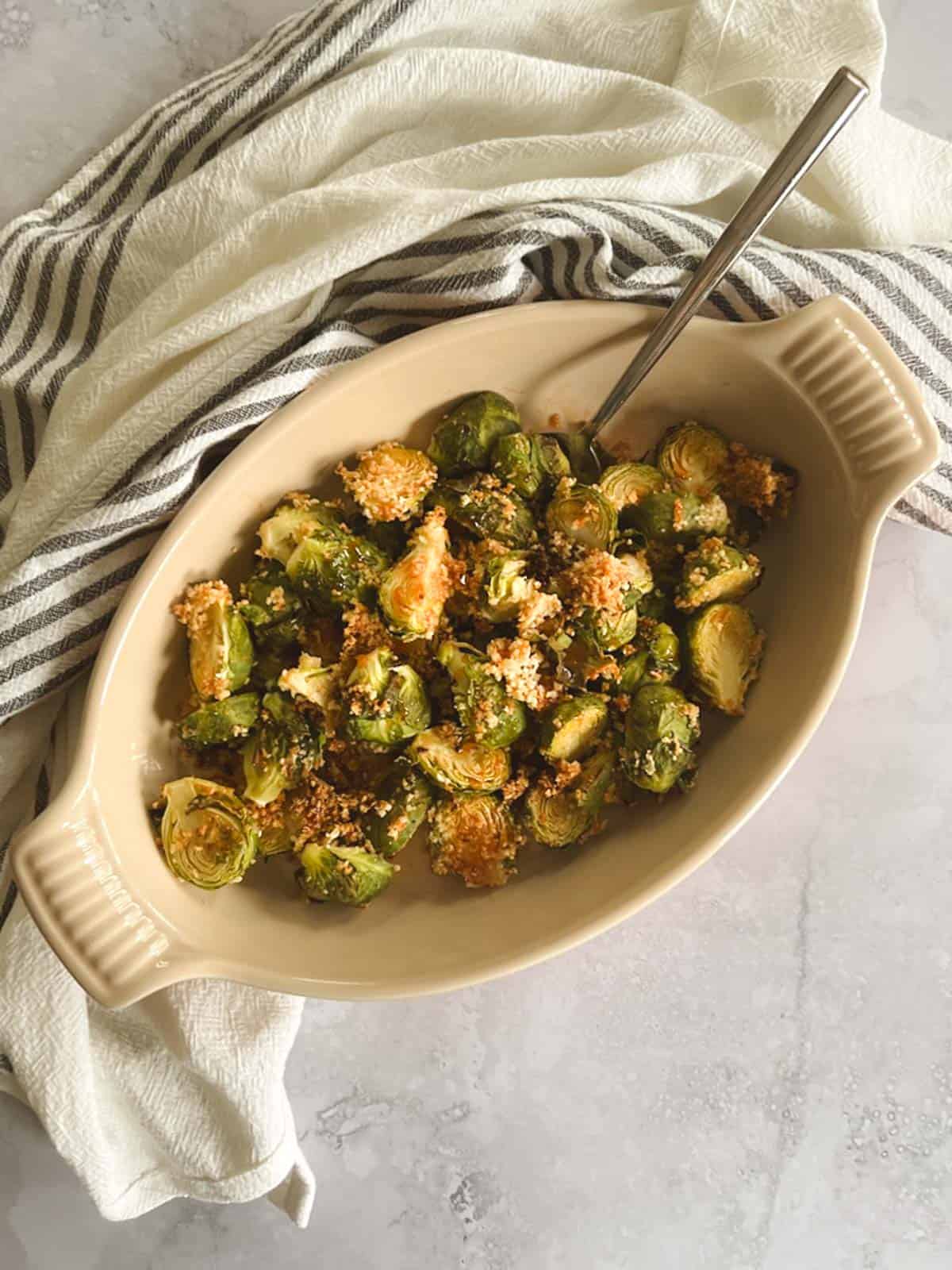 serving dish of crispy brussels sprouts with spicy maple syrup on a diagonal with a serving spoon and striped towel