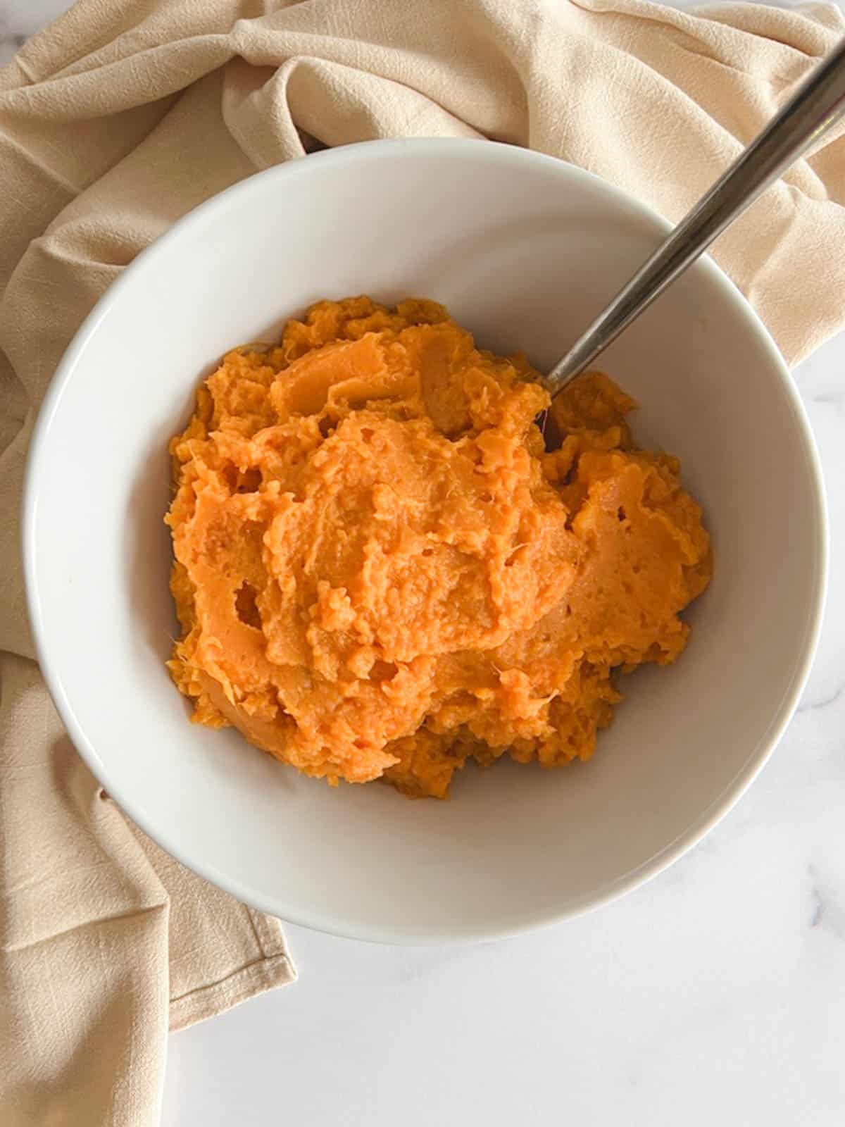 overhead view of white bowl containing instant pot mashed sweet potatoes with a serving spoon; beige towel in background