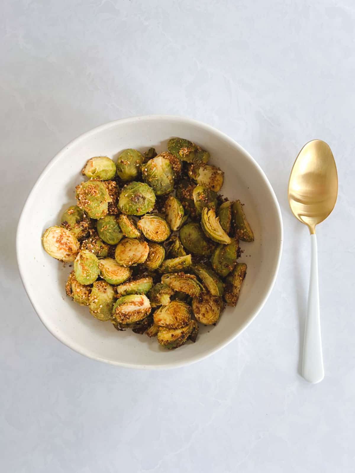 overhead view of white bowl containing crispy Brussels sprouts roasted in the oven with a gold and white serving spoon to its right