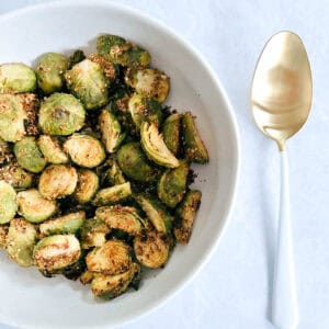 overhead of off-centered white bowl containing crispy brussels sprouts next to a gold and white serving spoon