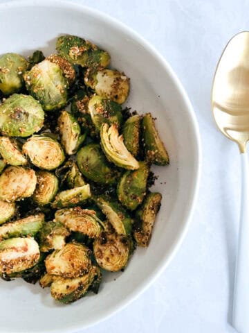 overhead of off-centered white bowl containing crispy brussels sprouts next to a gold and white serving spoon