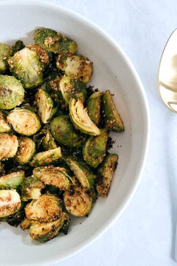 overhead of off-centered white bowl containing crispy brussels sprouts next to a gold and white serving spoon