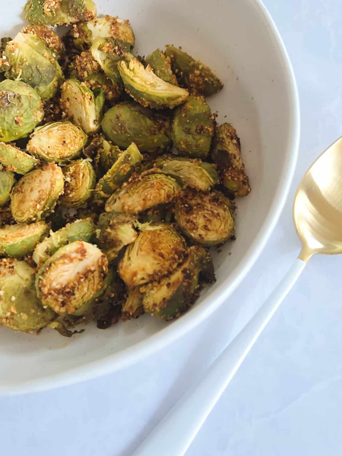 close up of part of a white bowl containing crispy Brussels sprouts roasted in the oven with a partial view of a gold and white serving spoon on a diagonal to the right