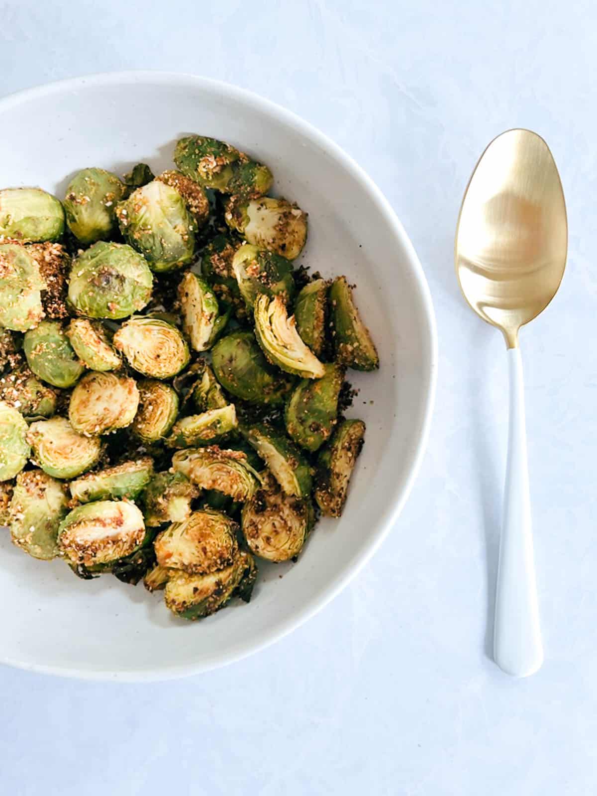 partial overhead view of white bowl containing crispy brussels sprouts roasted in the oven next to a gold and white serving spoon