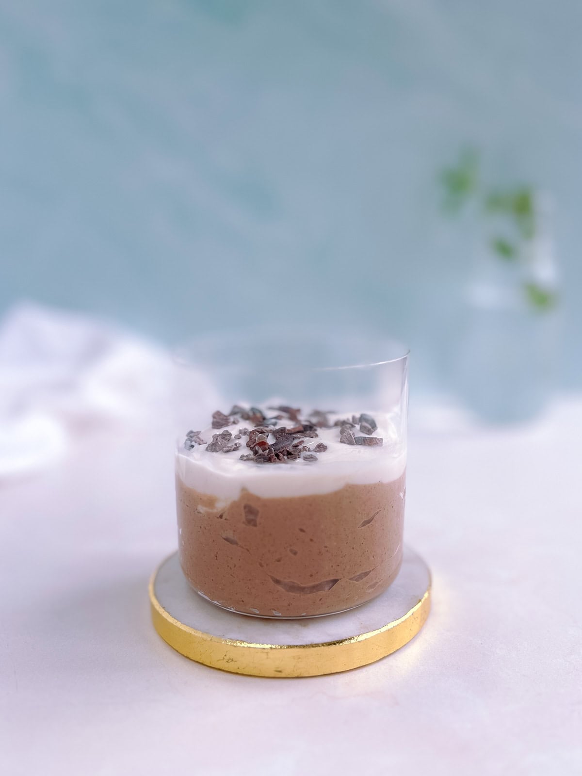 glass containing a serving of mint chocolate overnight oats garnished with vegan yogurt and cacao nibs; white napkin and plant in vase in the background