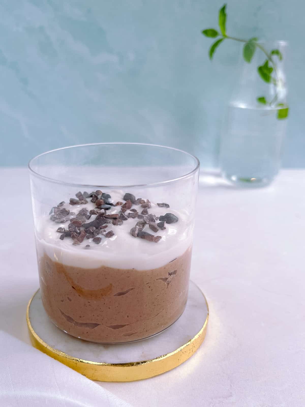 glass of mint chocolate overnight oats topped with vegan yogurt and garnished with cacao nibs, on a white and gold coaster with a glass vase in the background