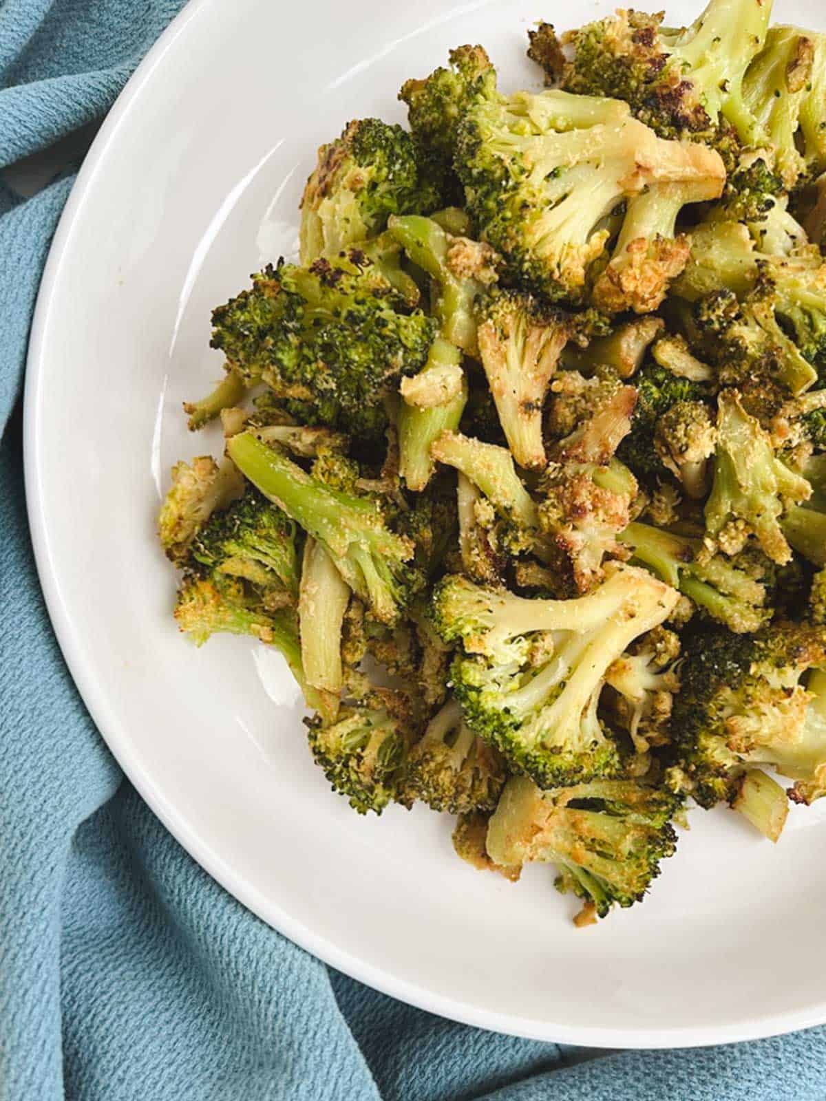 overhead partial view of white bowl containing oil-free roasted broccoli on a light blue kitchen towel