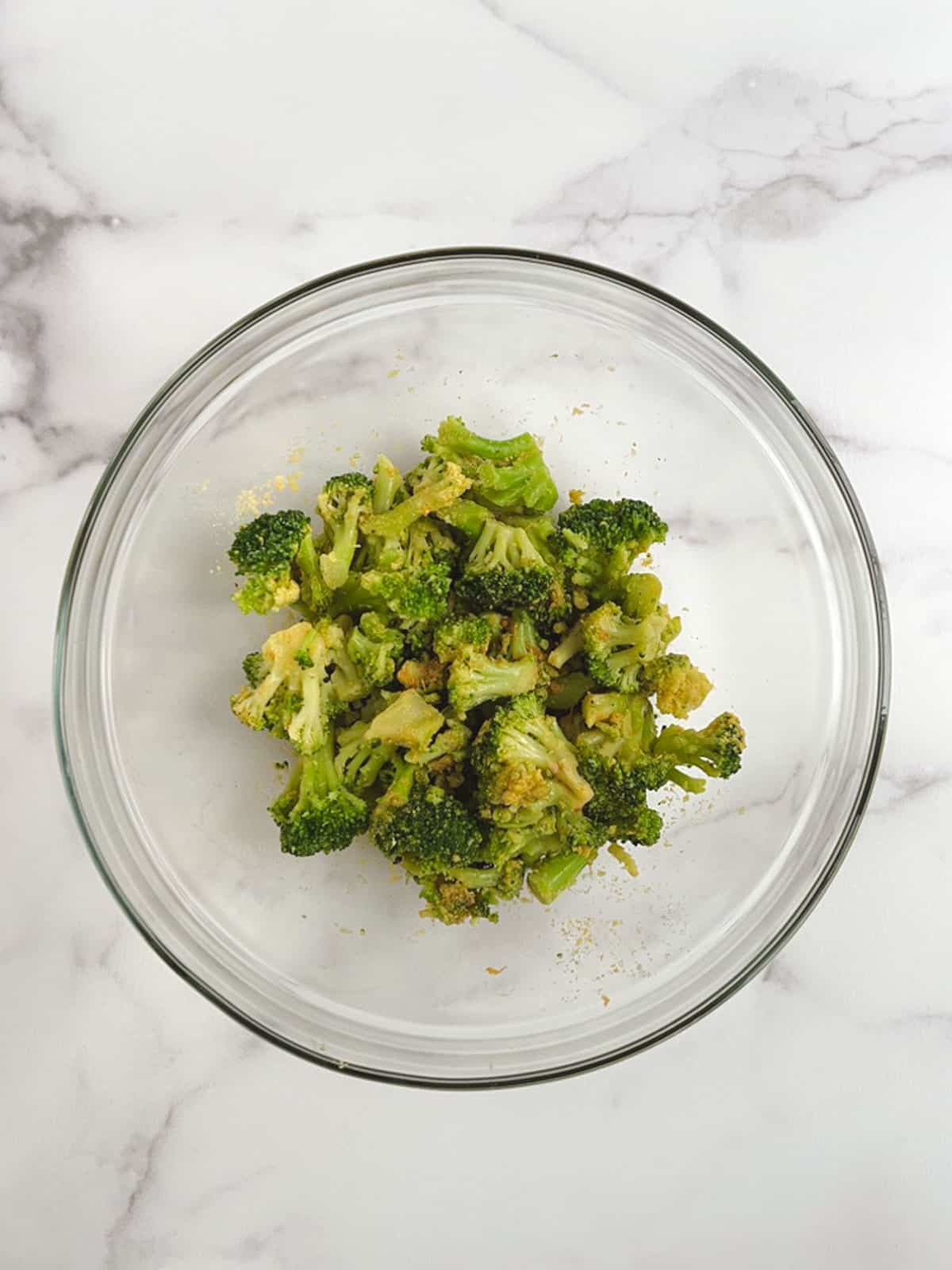 overhead view of glass bowl containing broccoli mixed with seasoning, on a white marble countertop