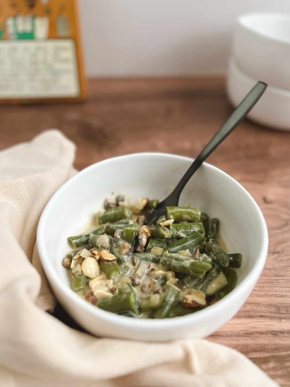 closeup of small white bowl containing serving of vegan green bean casserole with a black fork; decorative wooden plaque and stacked white bowls blurred in the background