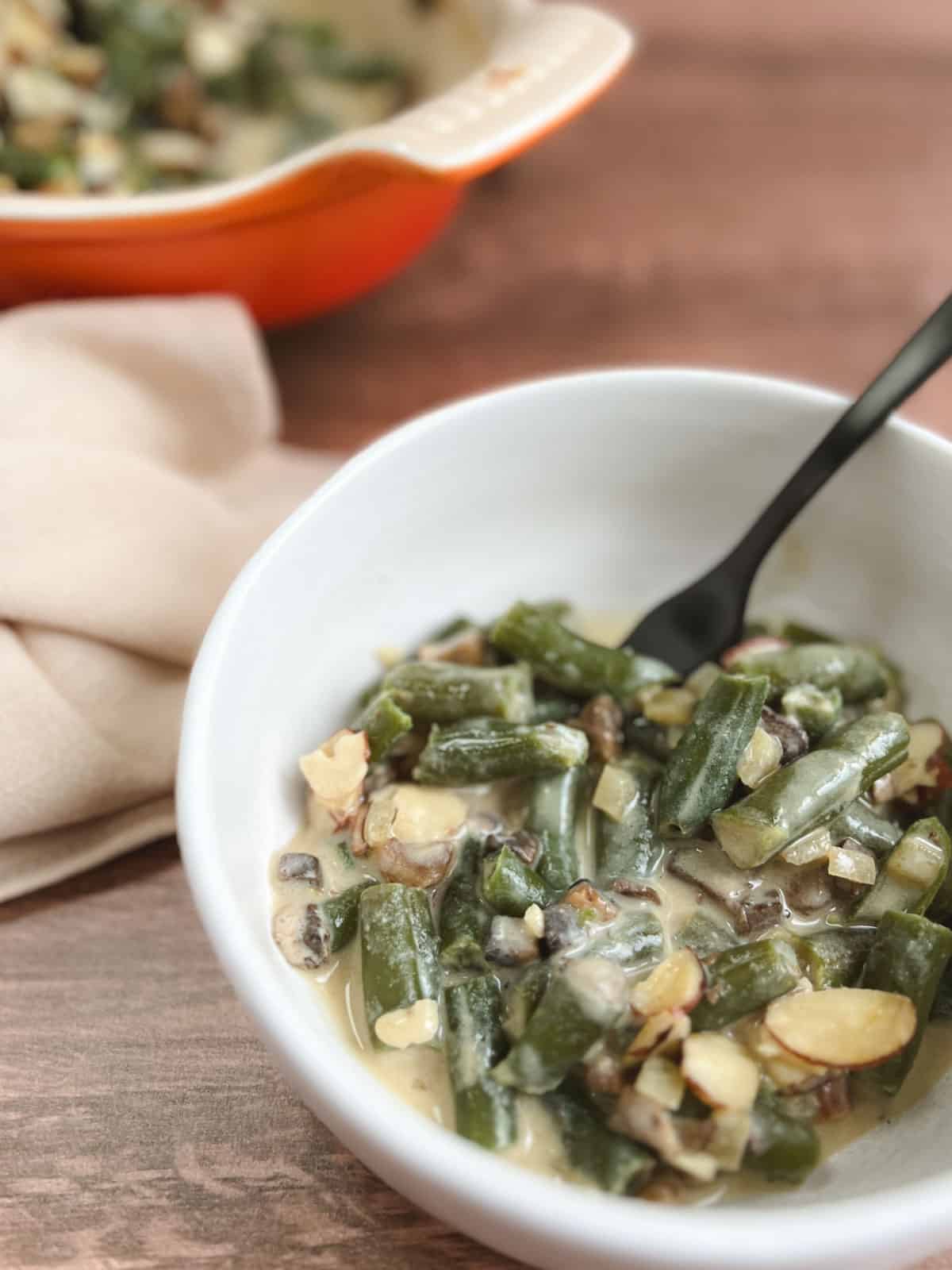 closeup of small white bowl containing serving of vegan green bean casserole with a blurred orange serving dish in the background