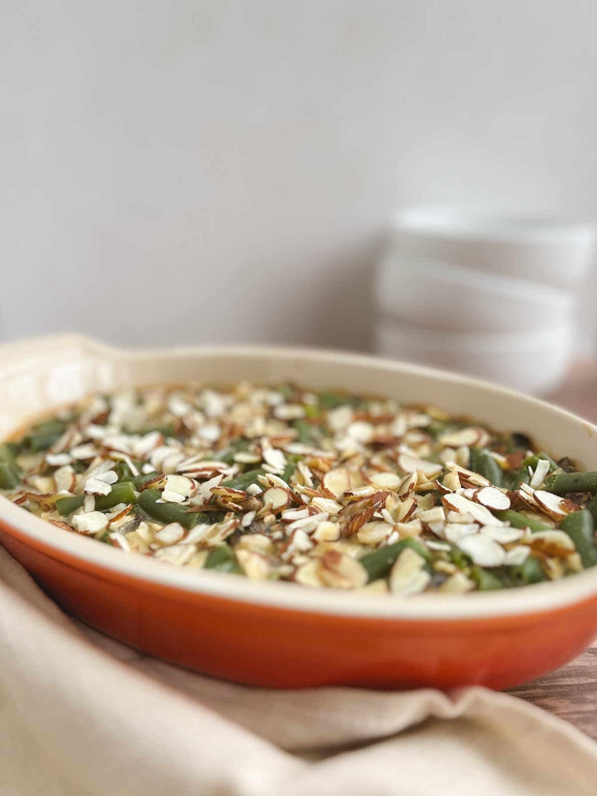 orange oval serving dish containing vegan green bean casserole with a beige napkin in the foreground and stacked white bowls blurred in the background