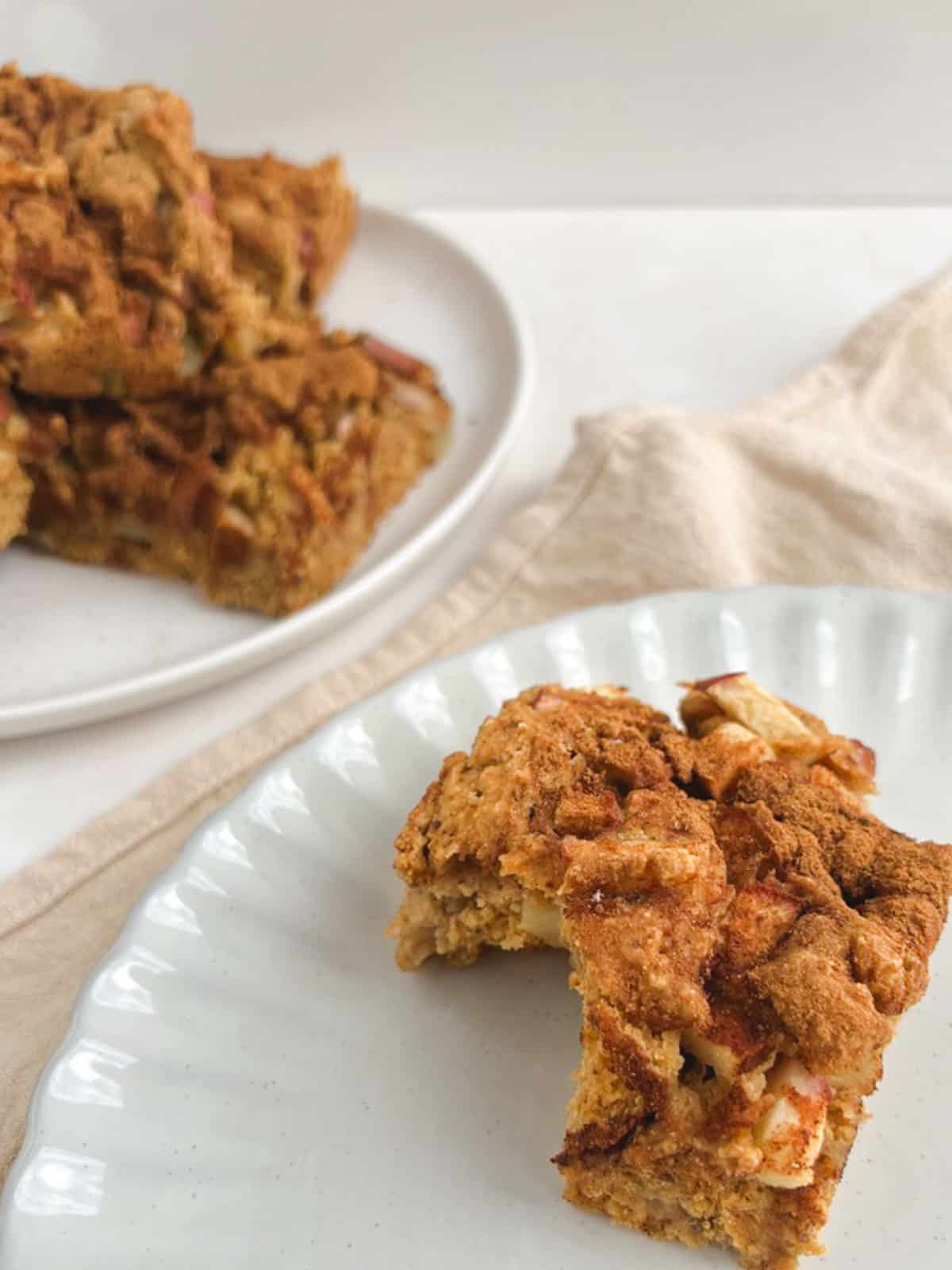 closeup of a square of apple coffee cake with a bite taken out of it on a small decorative plate with a large plate of apple coffee cake blurred in the background