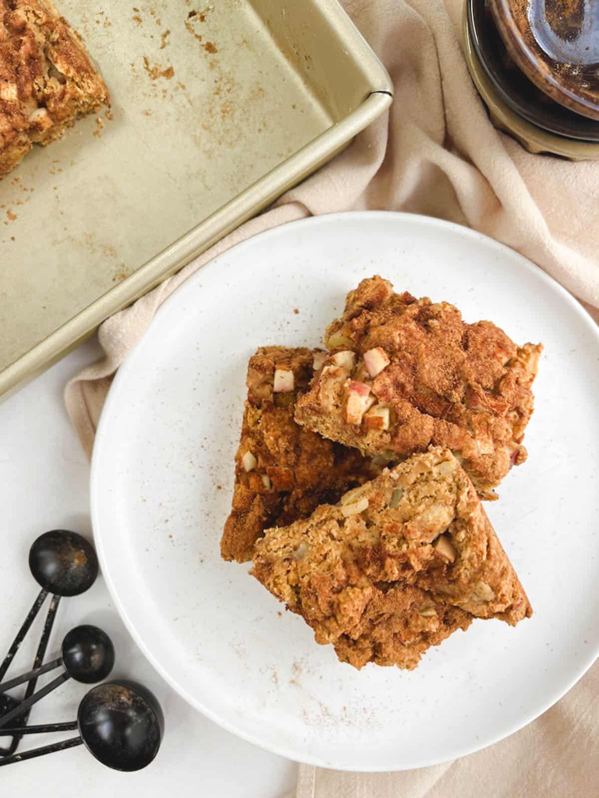 overhead view of three squares of apple coffee cake piled on a white plate next to a partially empty gold baking pan and measuring bowls and spoons