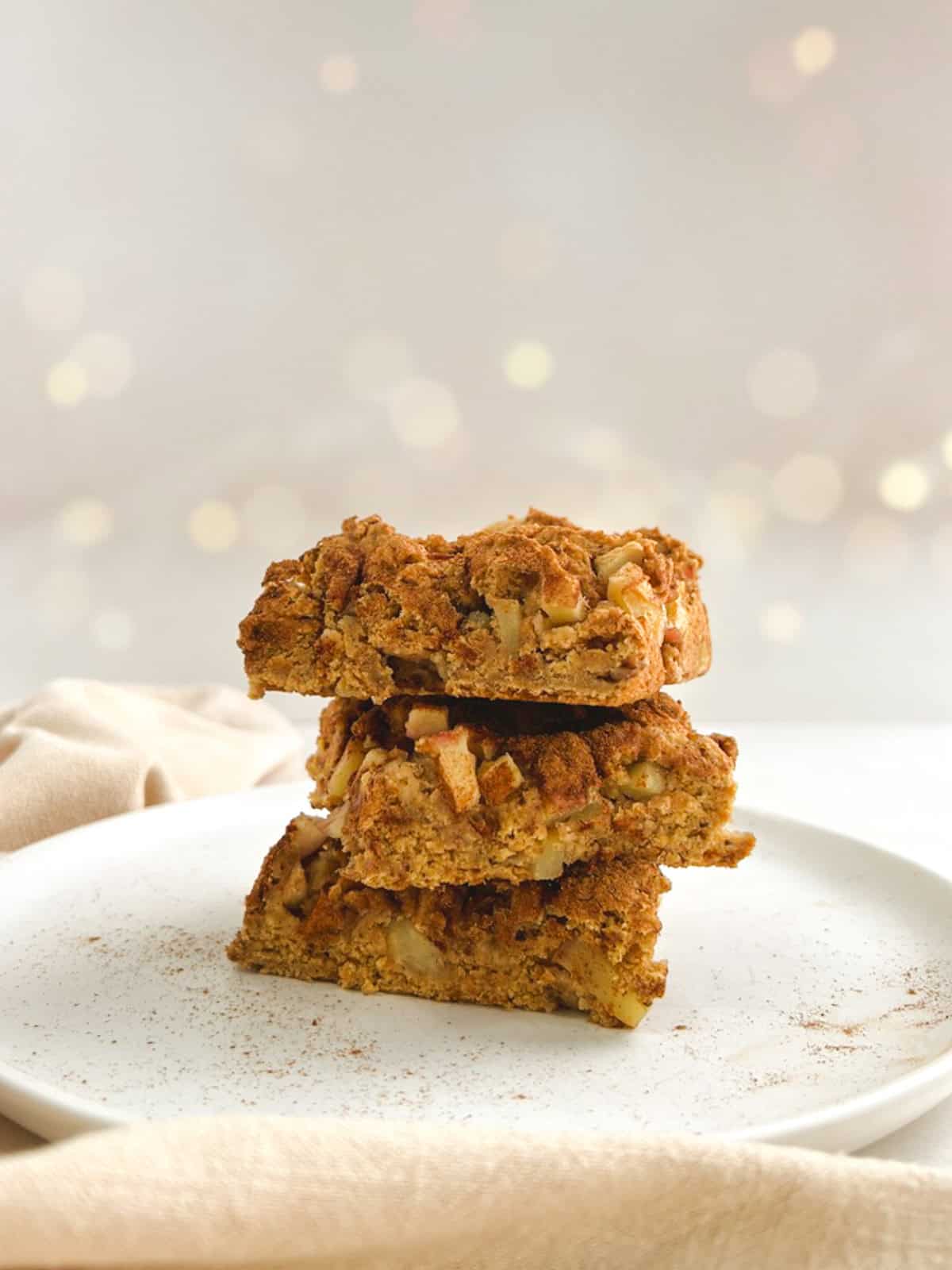 three squares of apple coffee cake piled on a white plate against a lightly glittering background