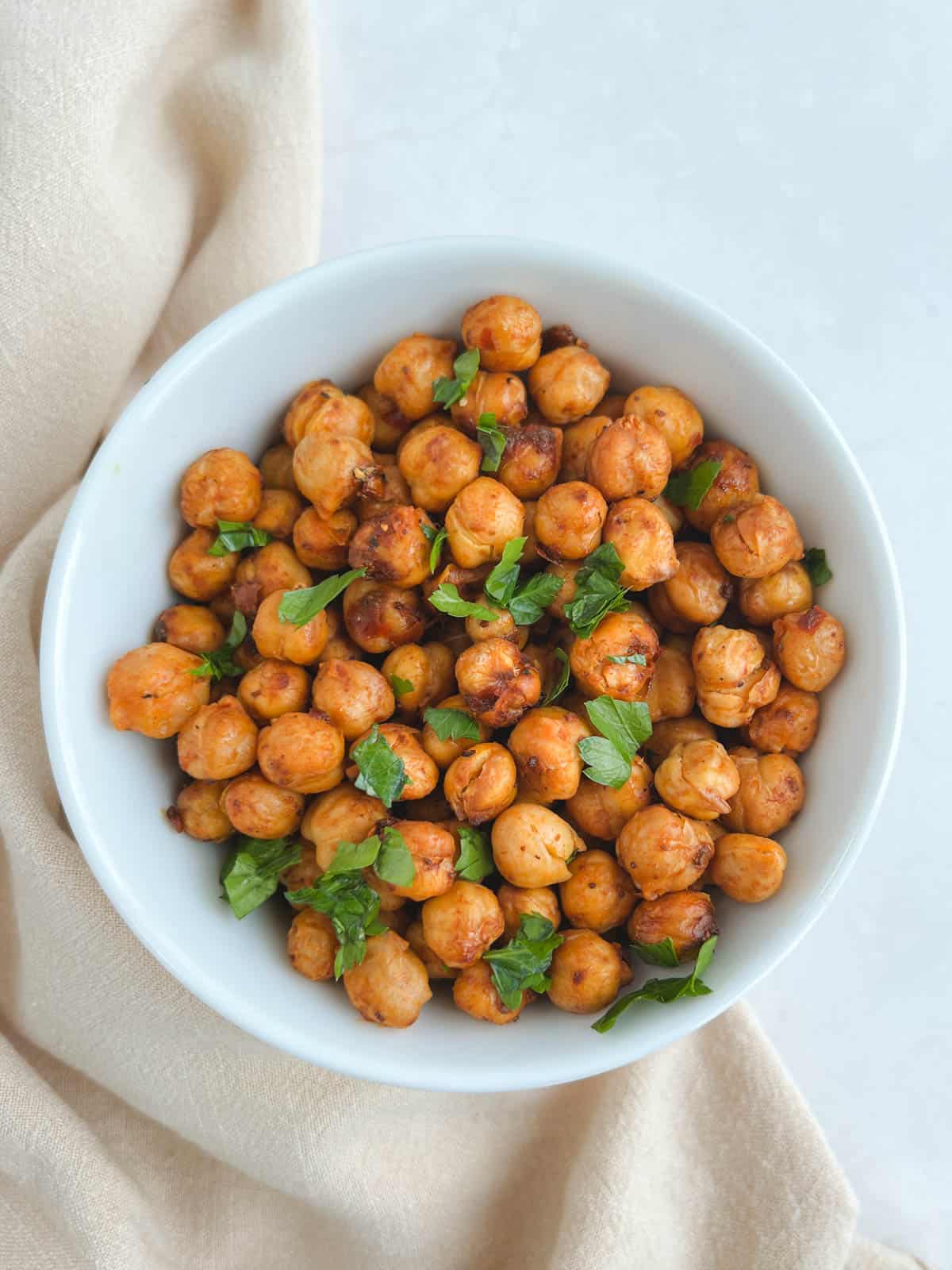 overhead view of white bowl containing harissa chickpeas garnished with parsley leaves on a white background with a cream-colored napkin