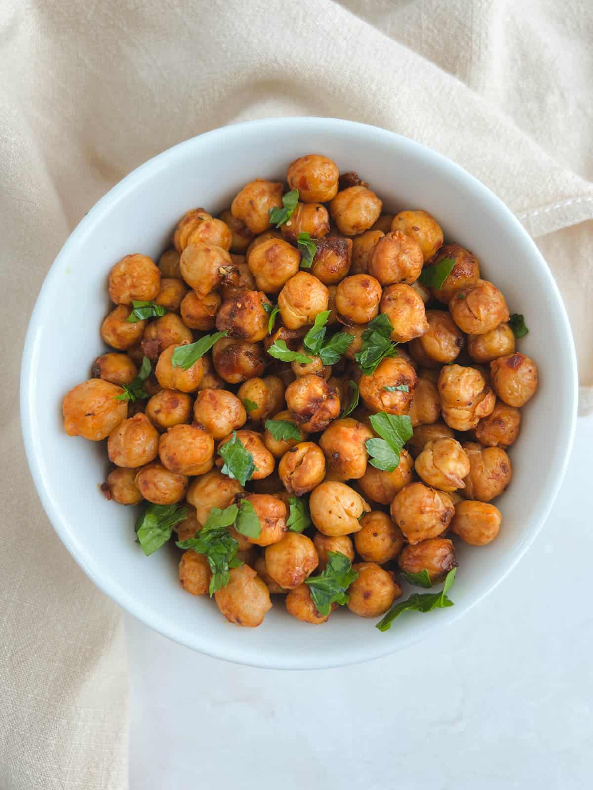 overhead view of harissa chickpeas in a white bowl on a cream-colored napkin