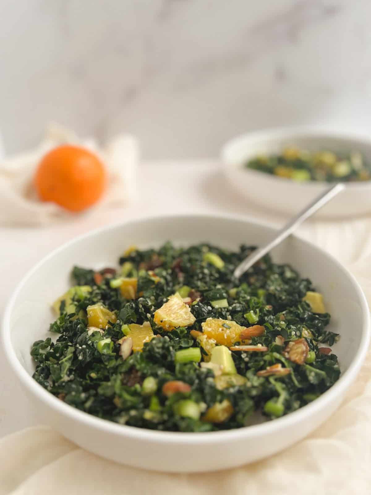 closeup of a small white bowl of kale citrus salad with an orange and another bowl of kale citrus salad blurred in the background