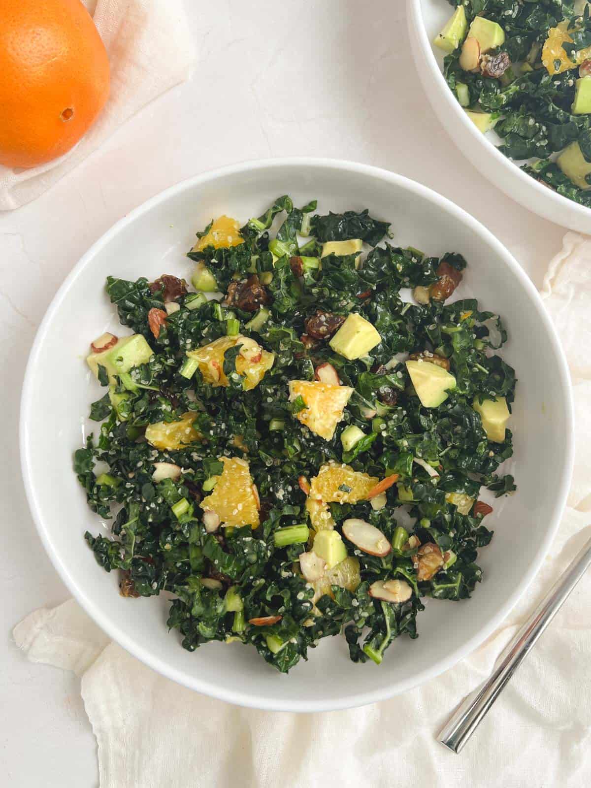 overhead view of a white bowl containing a serving of kale citrus salad next to a partially visible orange and a partially visible bowl of kale citrus salad